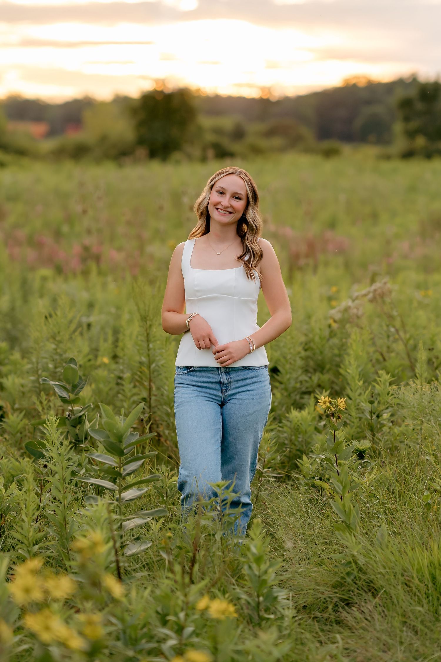 Young woman in white tank top and jeans smiling in a wildflower field at golden hour sunset.