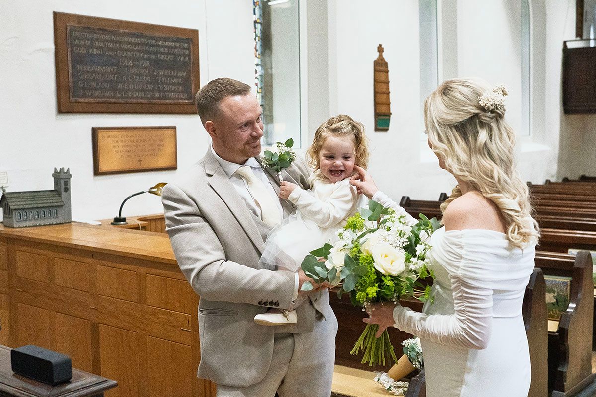 Bride and groom holding a young child during a church wedding ceremony, with the bride holding a white floral bouquet.