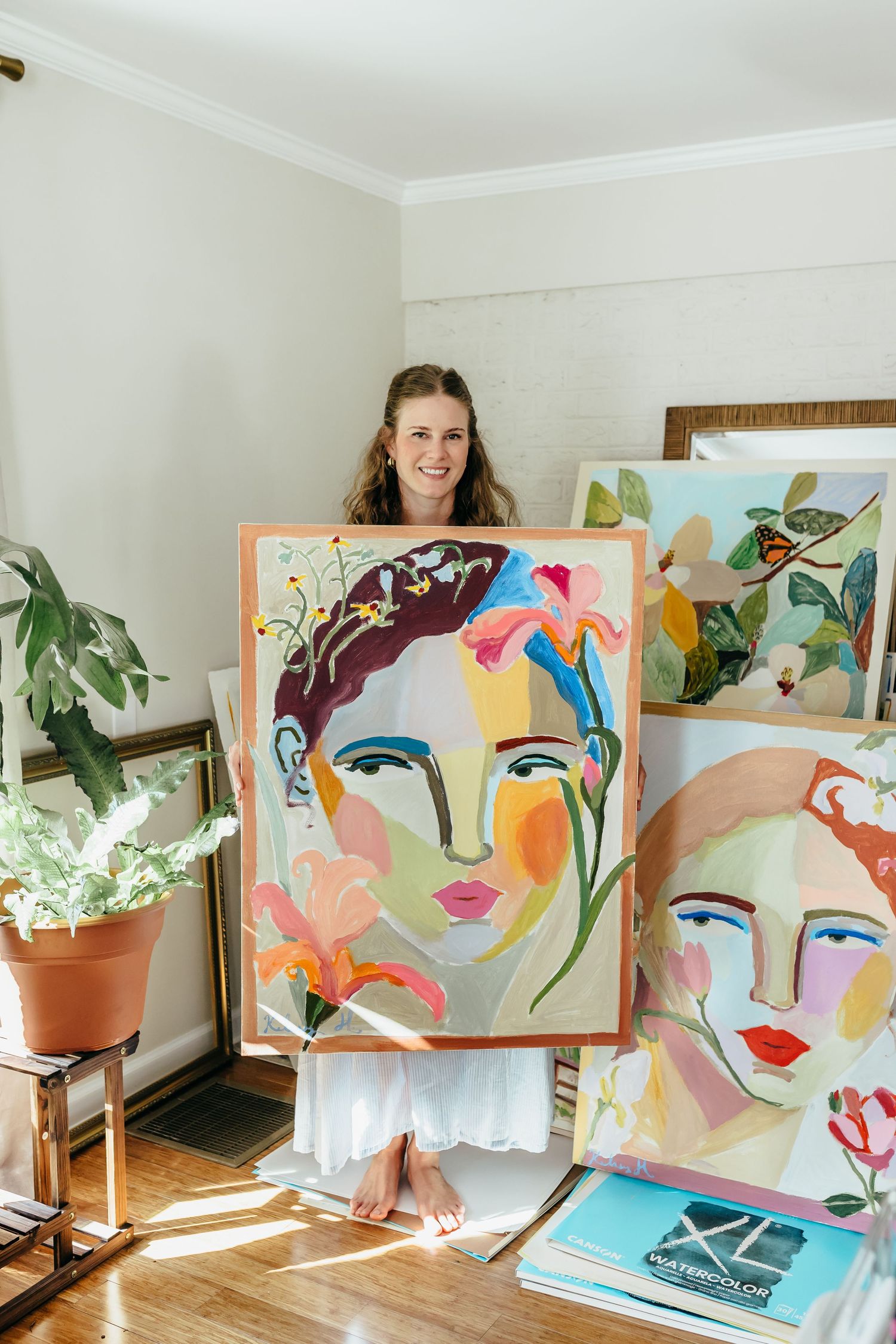 Artist smiling and holding colorful abstract face painting, surrounded by more artwork and plants in a bright studio.