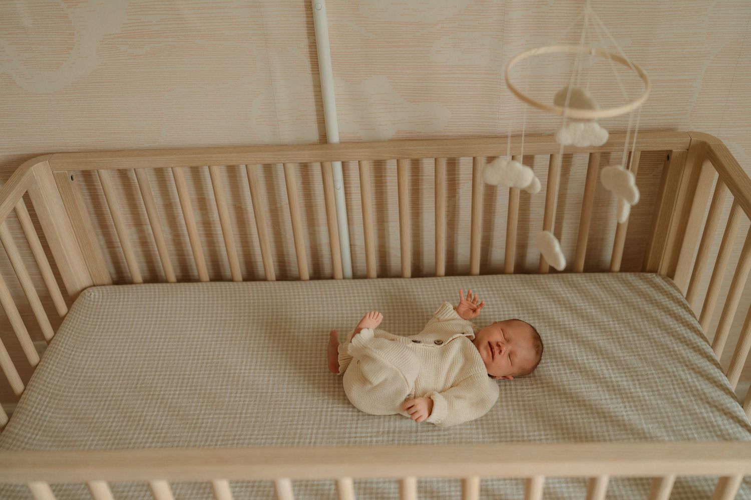 Newborn baby lying in a wooden crib with a mobile above, dressed in a white onesie on a sage green sheet.