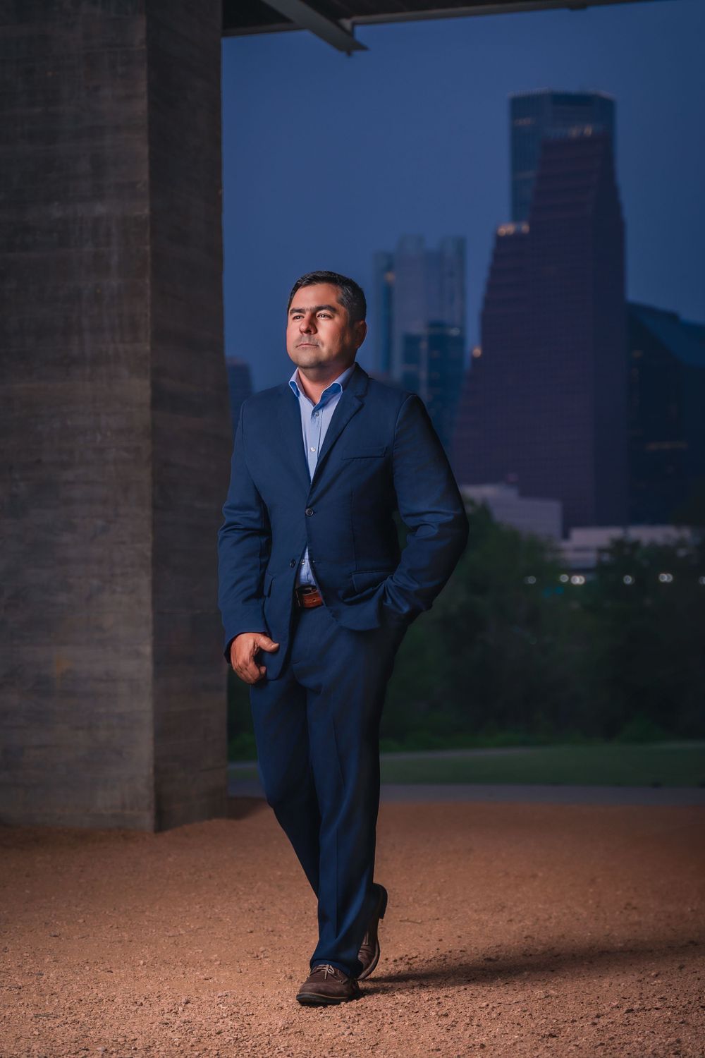 Professional in navy suit poses confidently with Houston skyline at dusk.