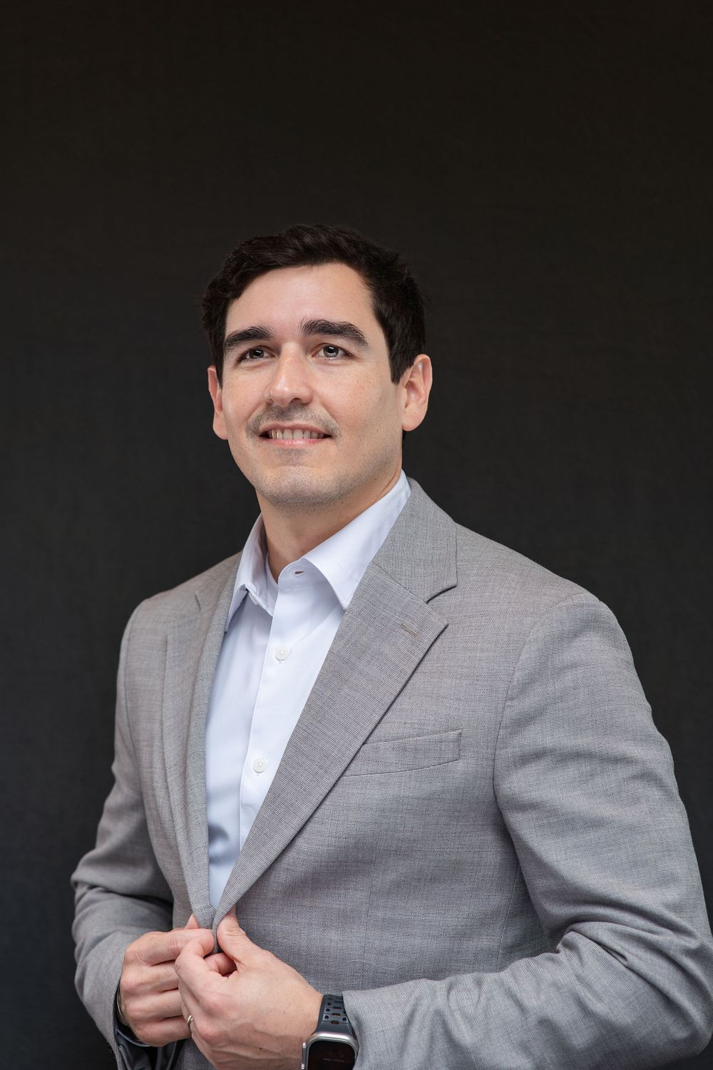 Professional man in gray blazer smiling confidently against dark background, wearing a smartwatch.