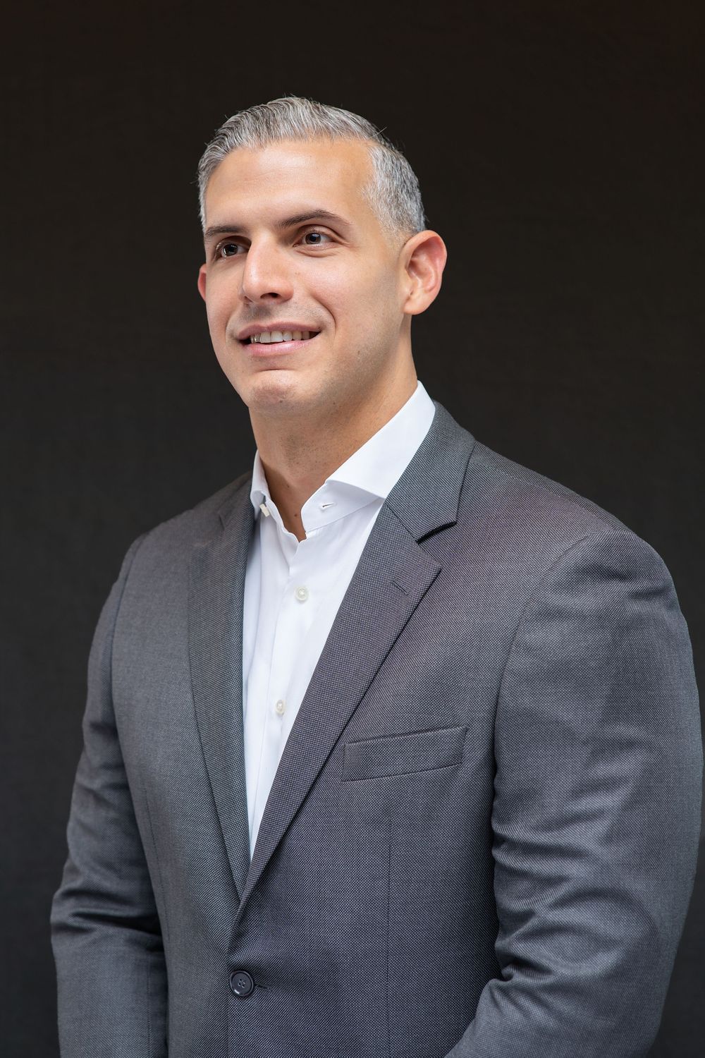 Professional man in gray suit and white shirt smiling confidently against dark background.