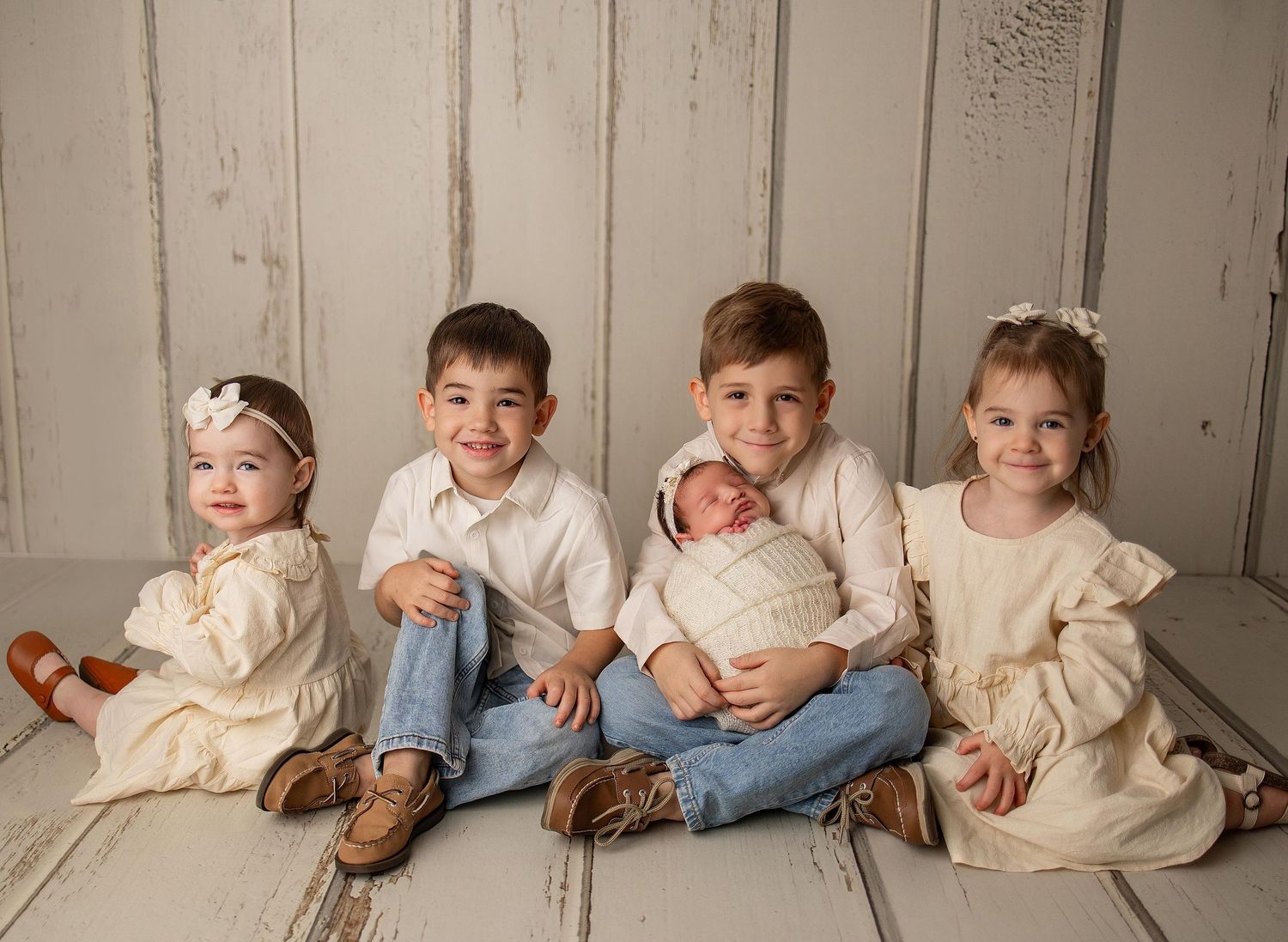 Four young siblings pose together on a white wooden floor, with the second oldest boy gently holding a swaddled newborn baby.