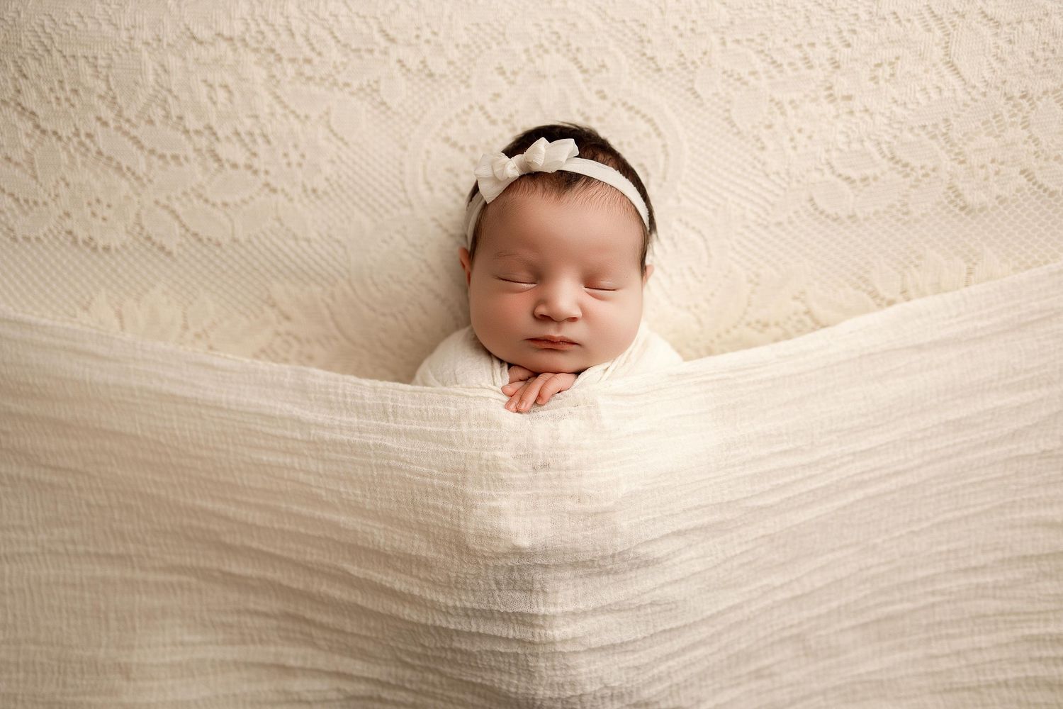 Peaceful newborn baby girl swaddled in cream wrap with floral headband, sleeping on white lace backdrop.
