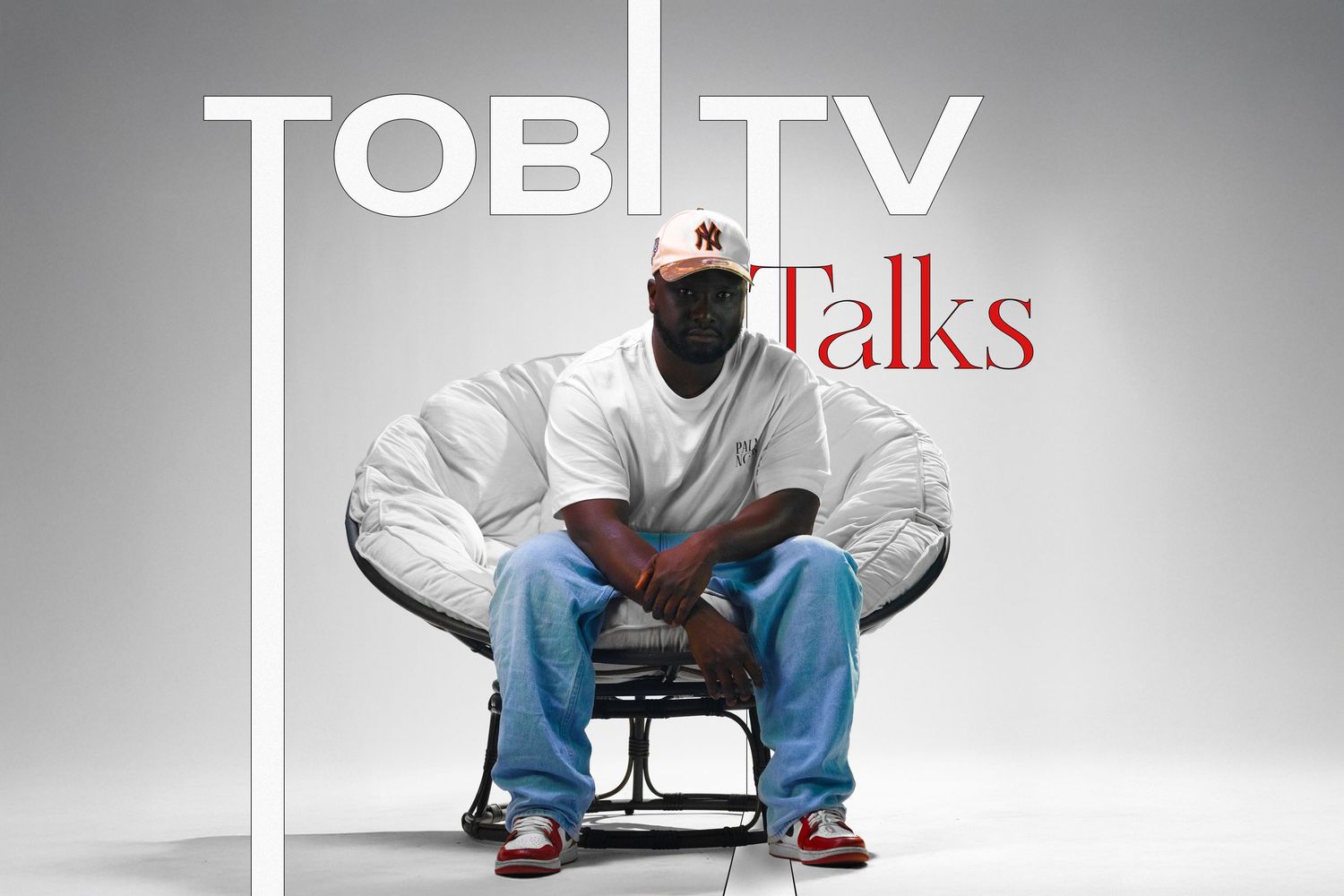 Man sitting in a chair posing for Tobi TV Talks promotional image wearing a white jacket and baseball cap.
