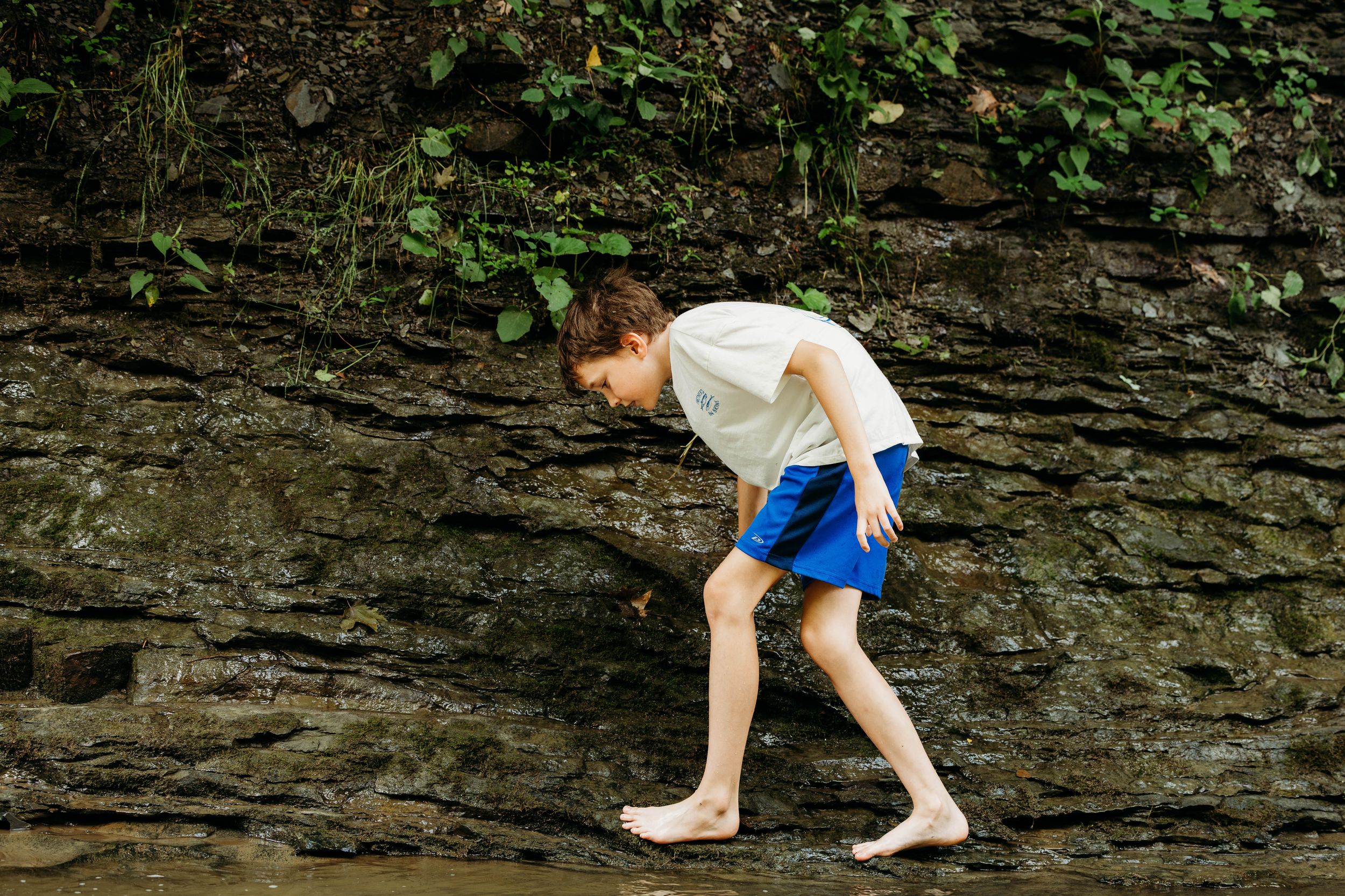 A barefoot boy in blue shorts explores wet rocks near a mossy stone wall outdoors.
