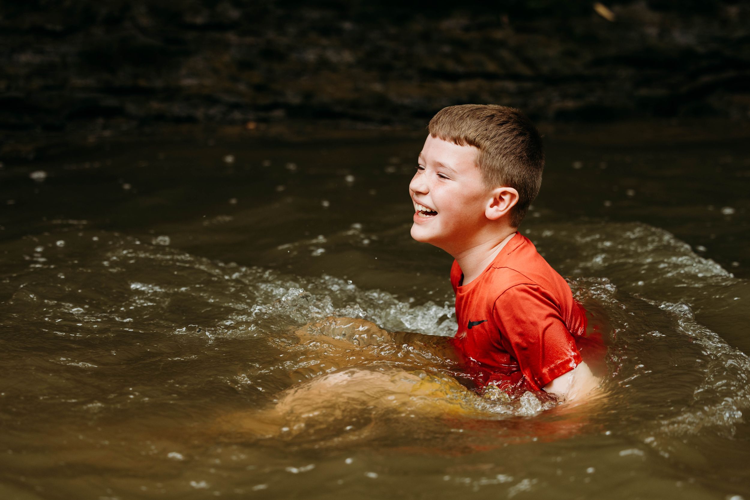 Happy boy in red shirt splashing and playing in a shallow creek, laughing with joy outdoors.