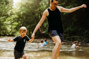 Woman and young boy playing in a shallow creek surrounded by lush green trees on a sunny day.