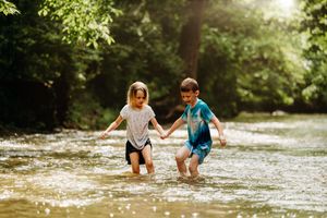 Two children hold hands and splash through a shallow sunlit creek surrounded by lush green trees.