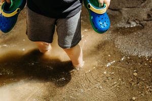 Child splashing barefoot in a muddy puddle while holding blue and yellow Crocs shoes outdoors.