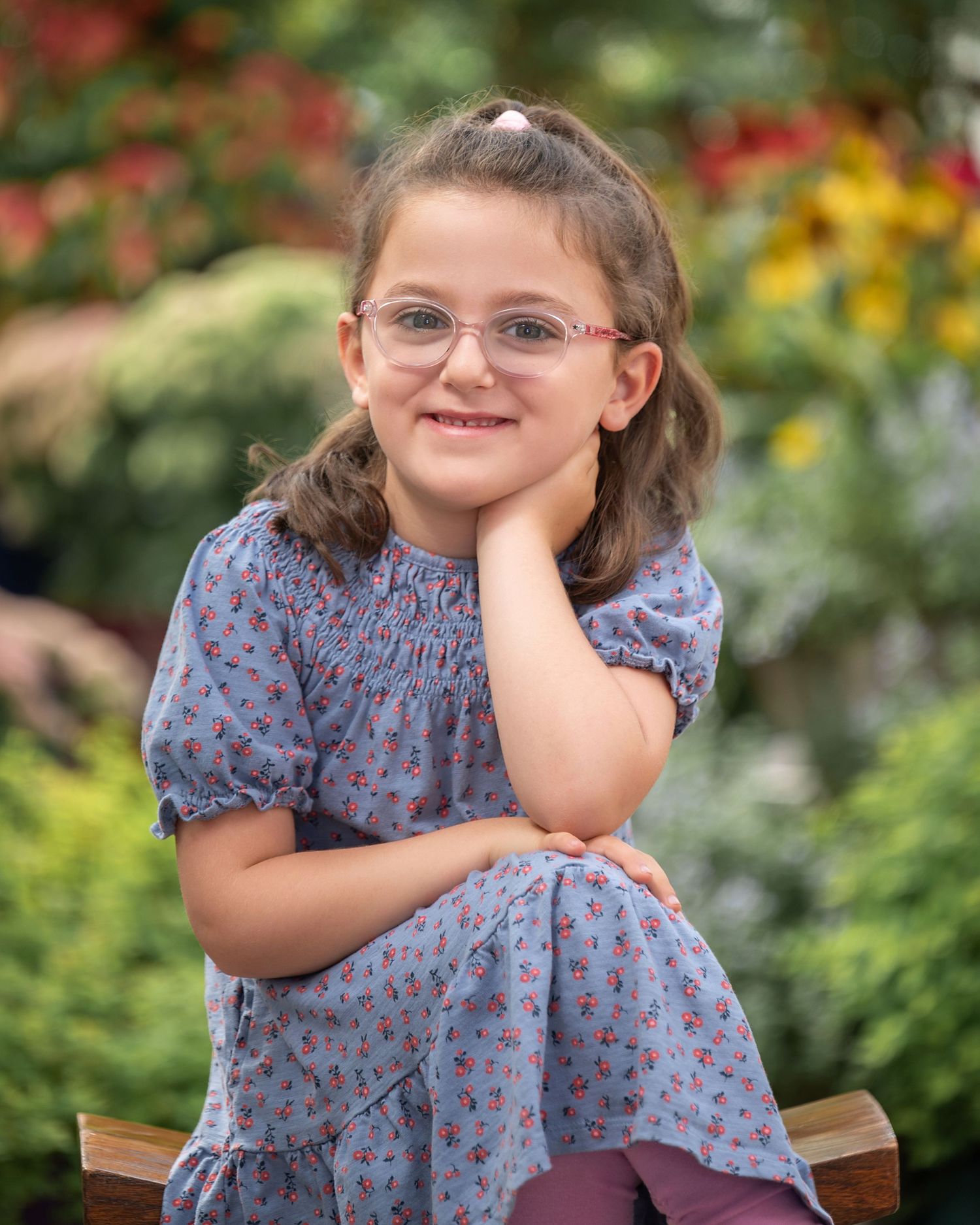 Young girl with glasses in floral blue dress sits smiling on bench in colorful garden setting.