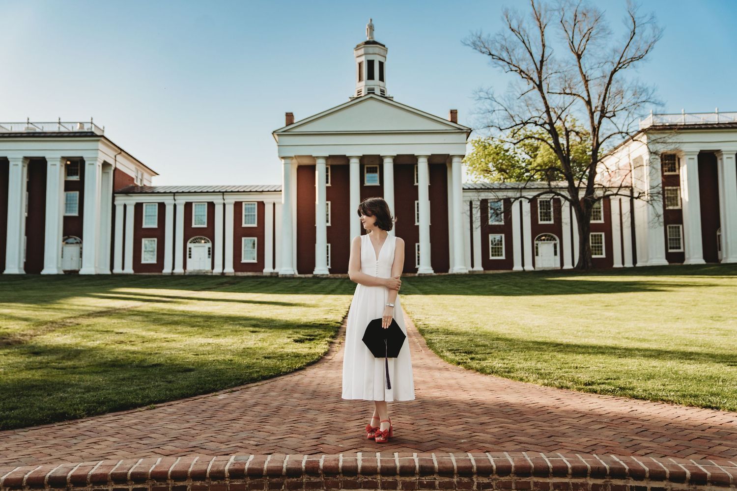 Young woman in white dress holding graduation cap stands on brick path before a classic red and white university building.