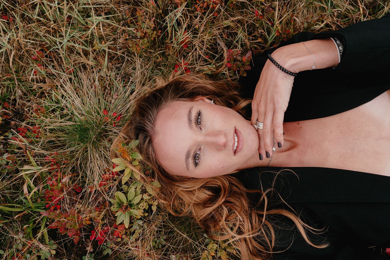 Woman with auburn hair lying in grass surrounded by red berries, wearing black top, gazing upward in serene outdoor portrait.