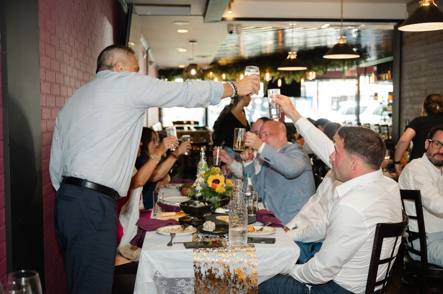 Group of people raising glasses in a toast at a wedding dinner celebration with floral centerpieces at Badageoni in Mt. Kisco, NY.