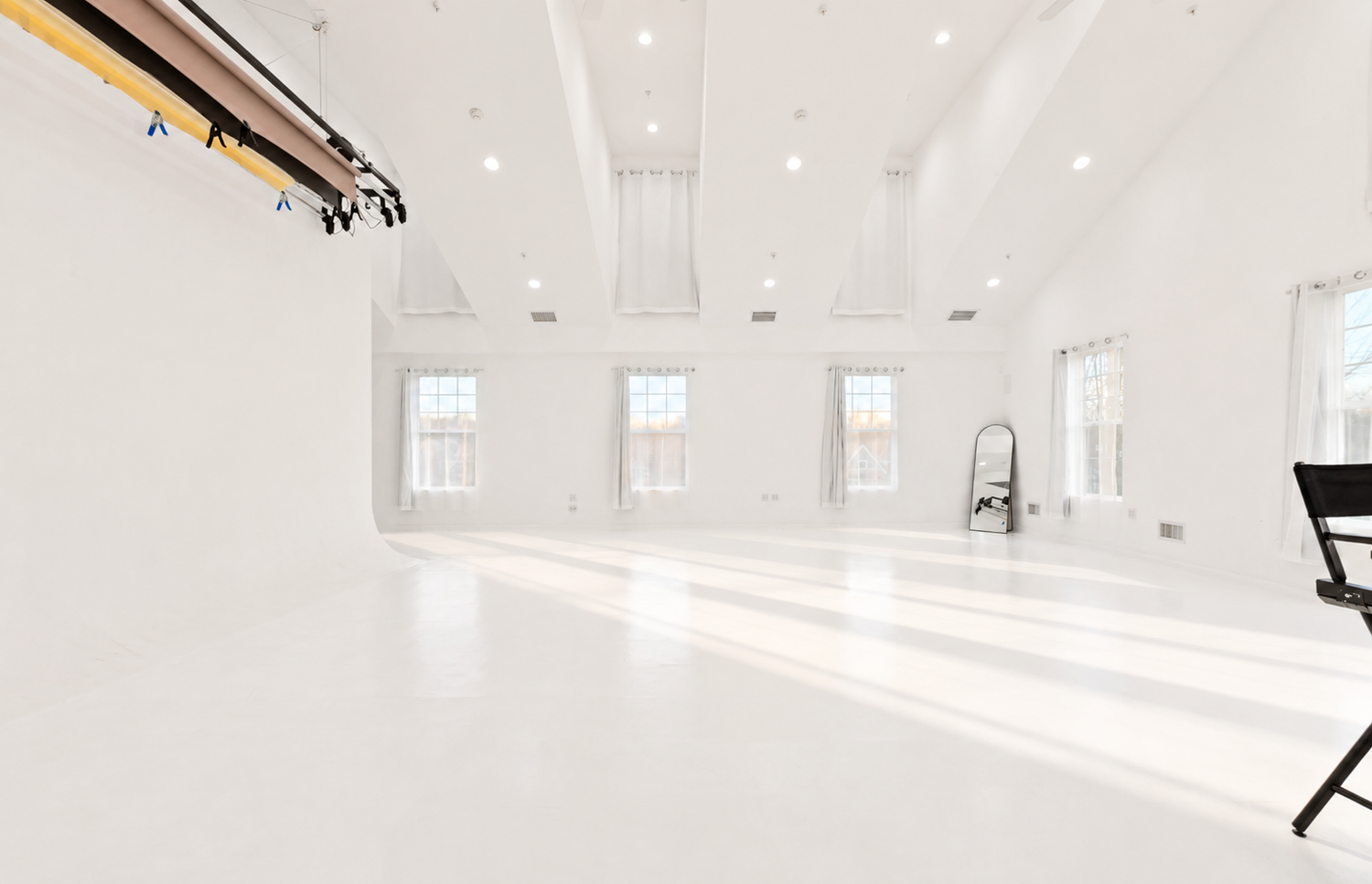 Bright white photography studio with high ceilings, large windows, seamless white floors, and hanging backdrop rails.