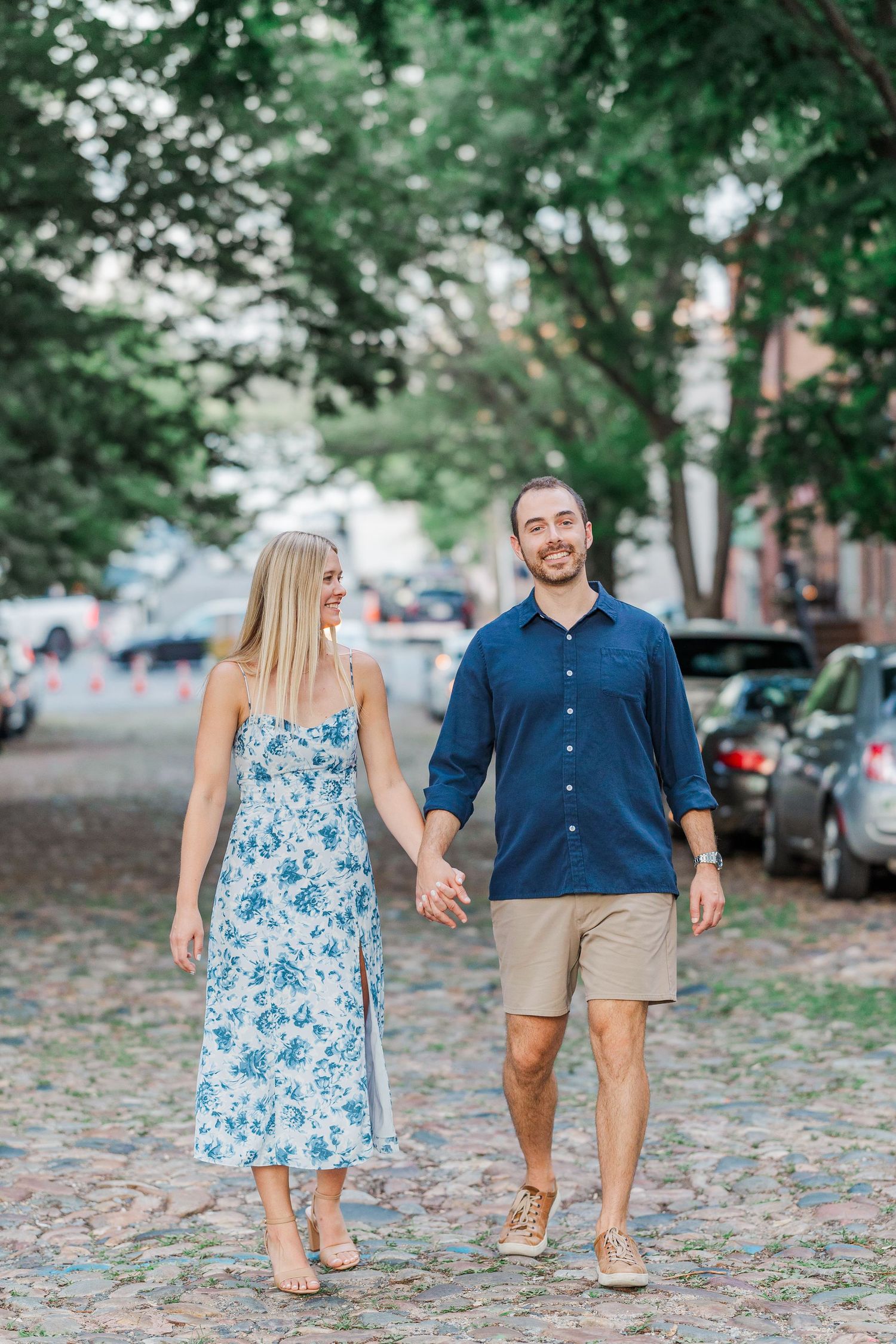 Couple holding hands walking on cobblestone street, woman in floral dress, man in navy shirt and khaki shorts.