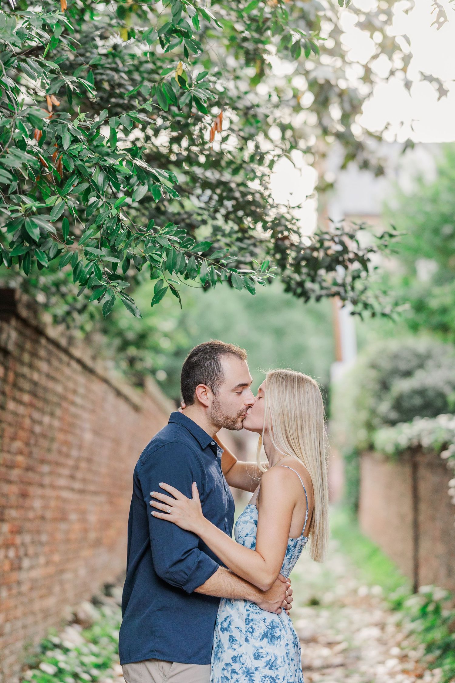 Couple sharing a romantic kiss outdoors, surrounded by lush green trees and a brick wall backdrop.