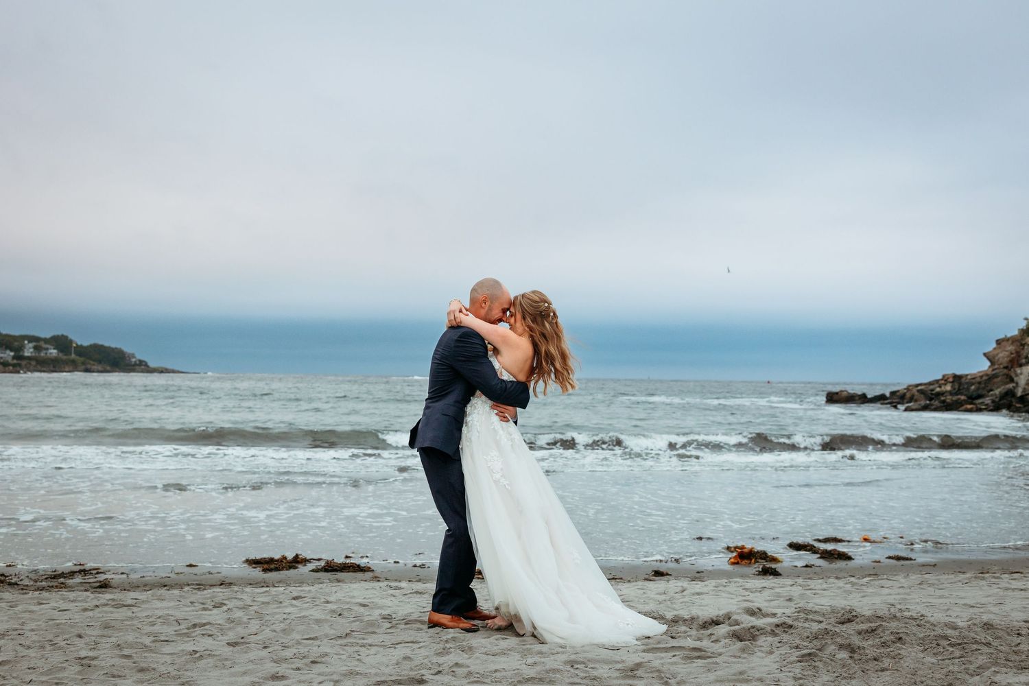 Couple sharing a romantic kiss on a sandy beach in wedding attire, with ocean waves and cloudy sky behind them.