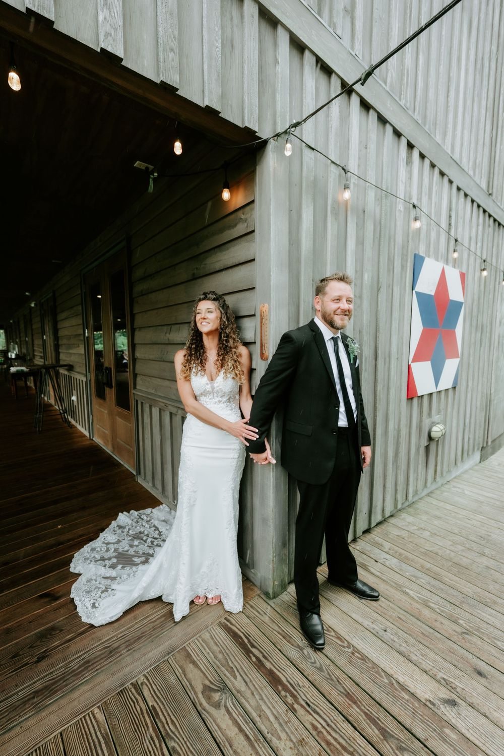 Bride and groom holding hands outside a rustic barn wedding venue with string lights and a decorative quilt.