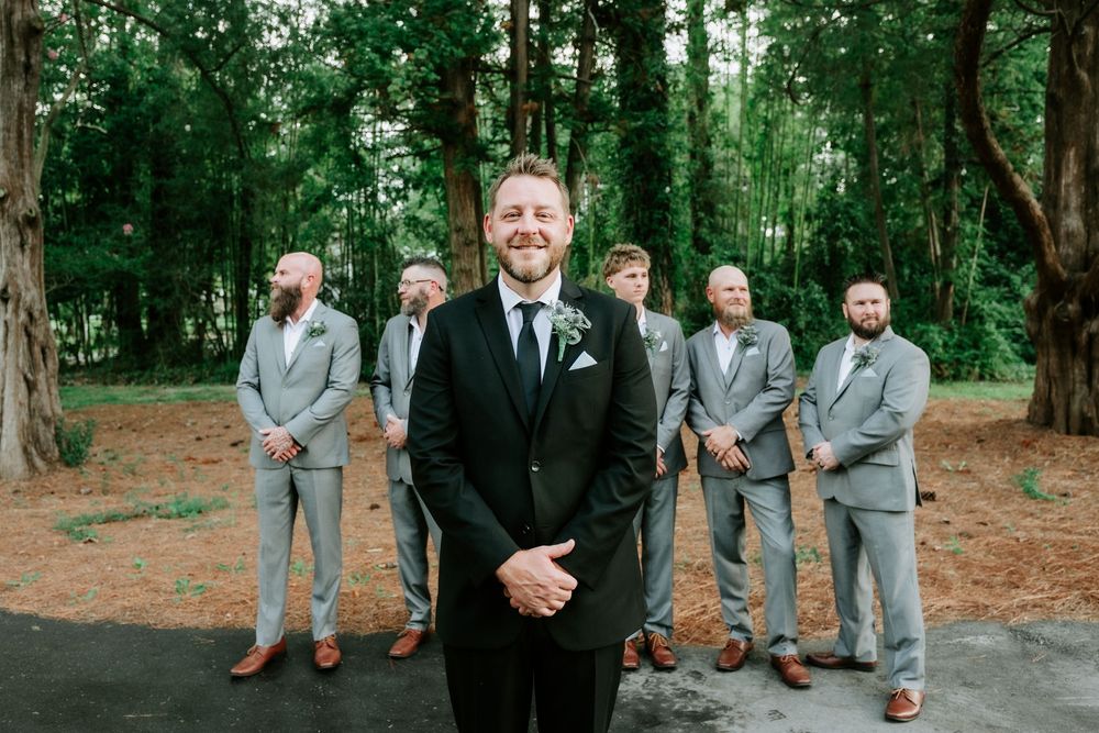 Groom in black suit stands before groomsmen in gray suits outdoors among tall green trees at a wedding.