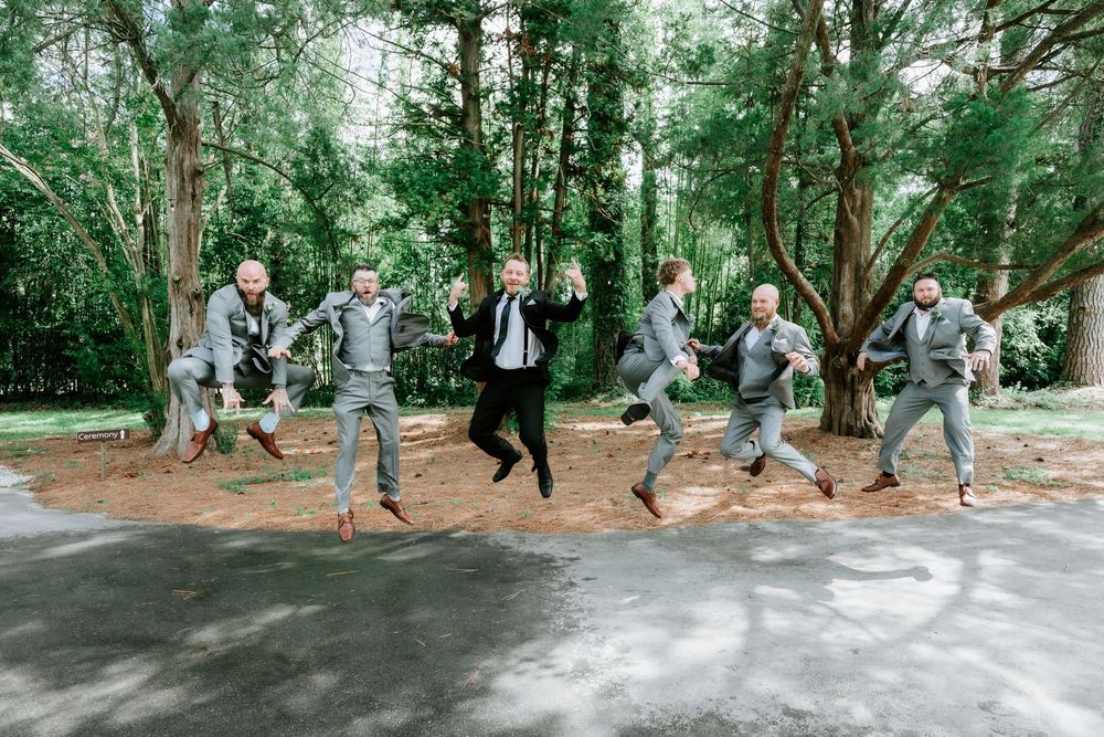 Wedding party in gray suits jumping joyfully on a path surrounded by lush green trees.