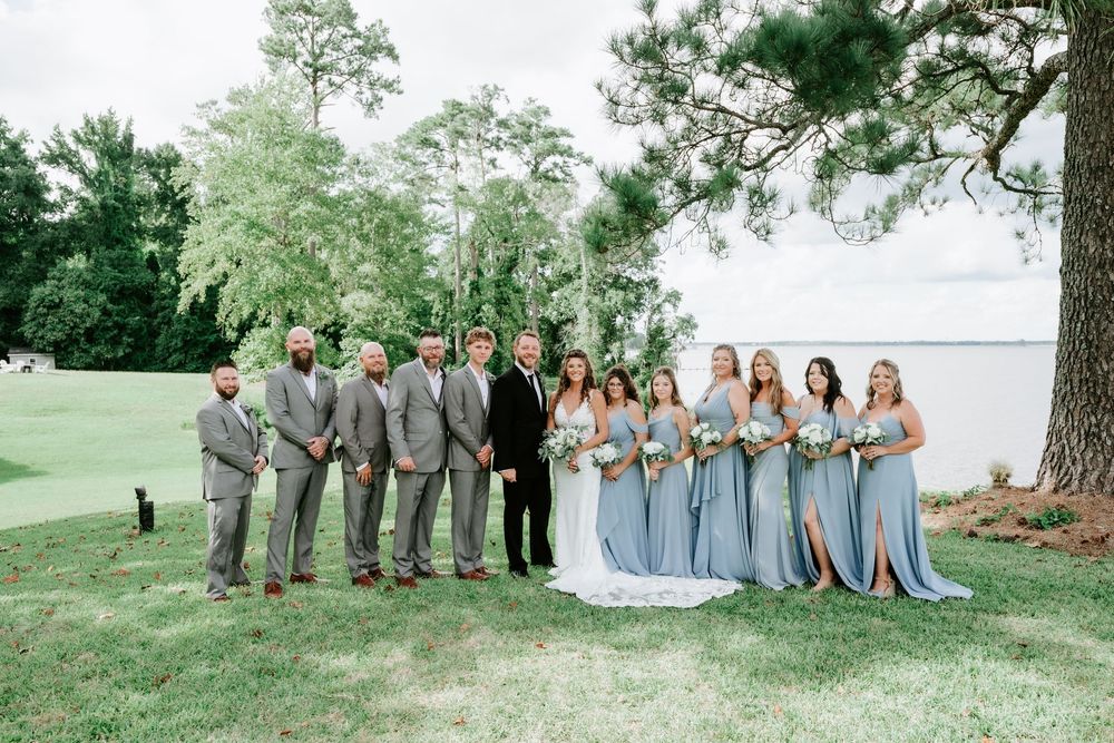 Wedding party poses outdoors near waterfront, bridesmaids in dusty blue gowns and groomsmen in gray suits flanking the bride and groom.