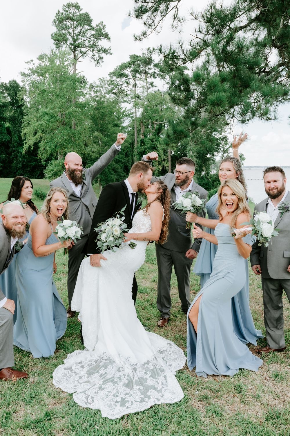 Joyful wedding party celebrates as groom lifts bride during outdoor photo session, surrounded by cheering bridesmaids and groomsmen.