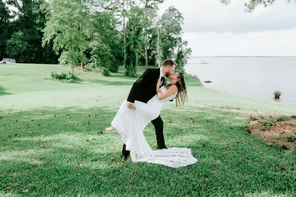 Groom dips bride for a romantic kiss on green lawn by waterfront during outdoor wedding photo session.