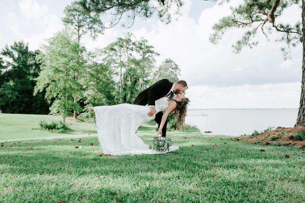 Groom dips bride in white wedding dress on lush green lawn near waterfront surrounded by trees.