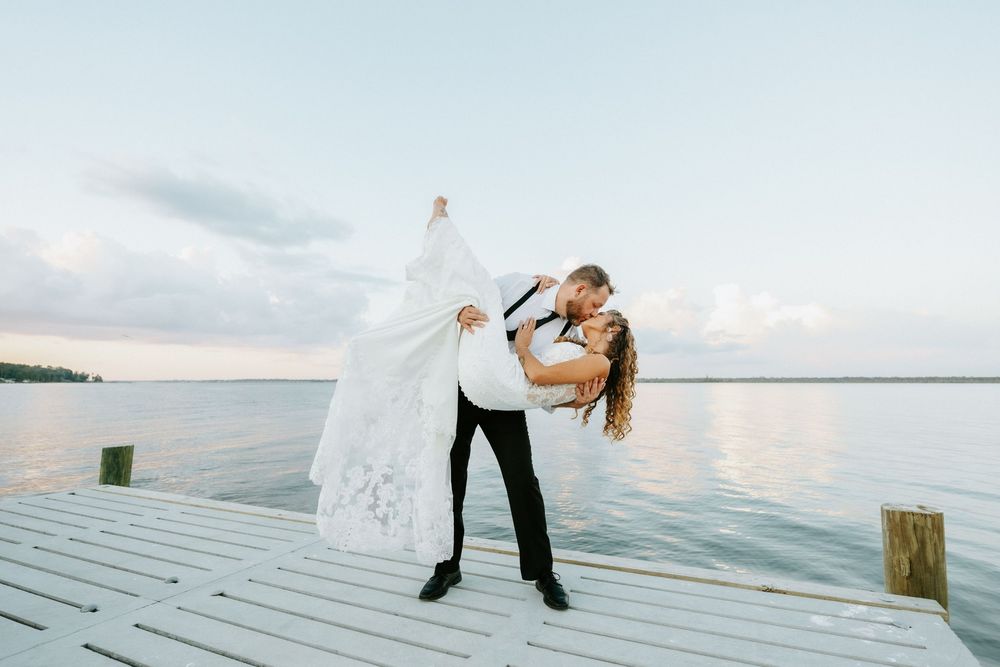 Bride and groom share a romantic dip kiss on a white dock overlooking calm water at sunset.