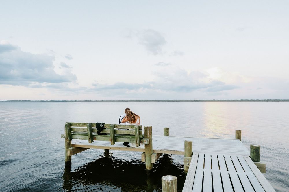 Couple sitting on a wooden dock bench overlooking a calm lake at sunset with cloudy skies.
