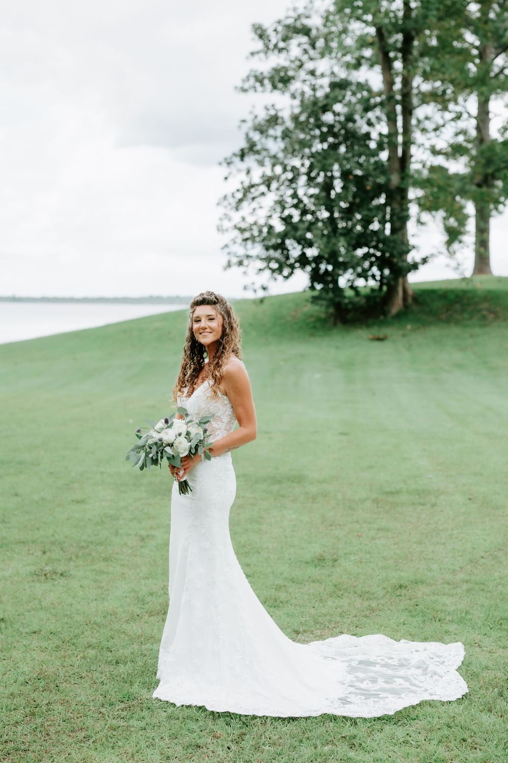 Bride in a lace mermaid gown holding a bouquet on a green lawn with a lake and trees in the background.