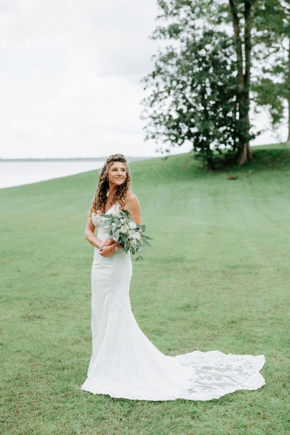 Bride in white lace gown with train holds bouquet on green lawn near water and trees.