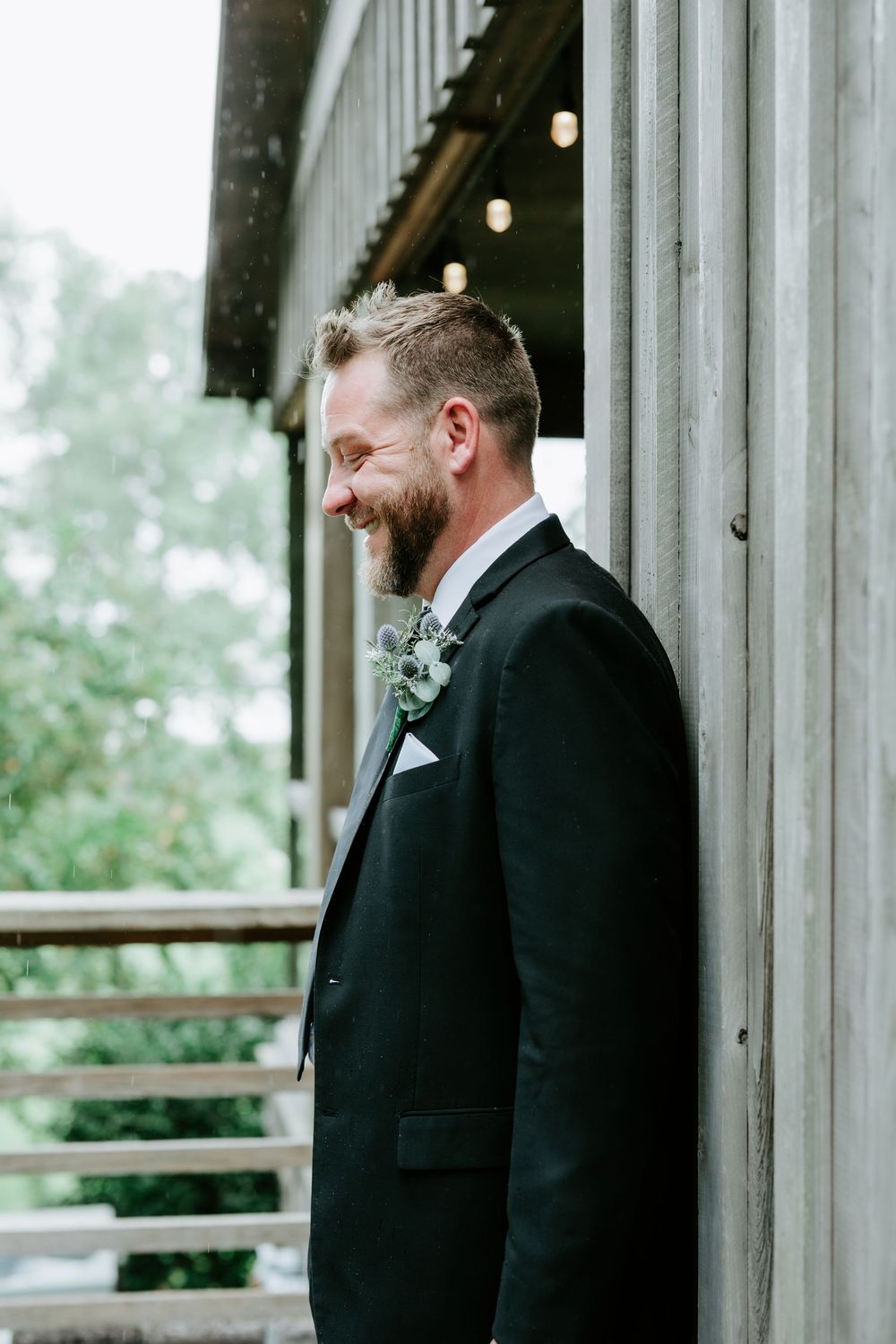 Smiling groom in black tuxedo with floral boutonniere leaning against rustic barn wall on wedding day.