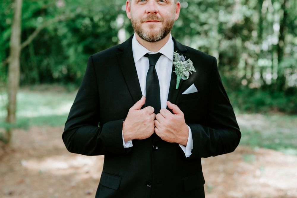 Groom in black suit with boutonniere adjusting jacket lapels outdoors at wedding ceremony.
