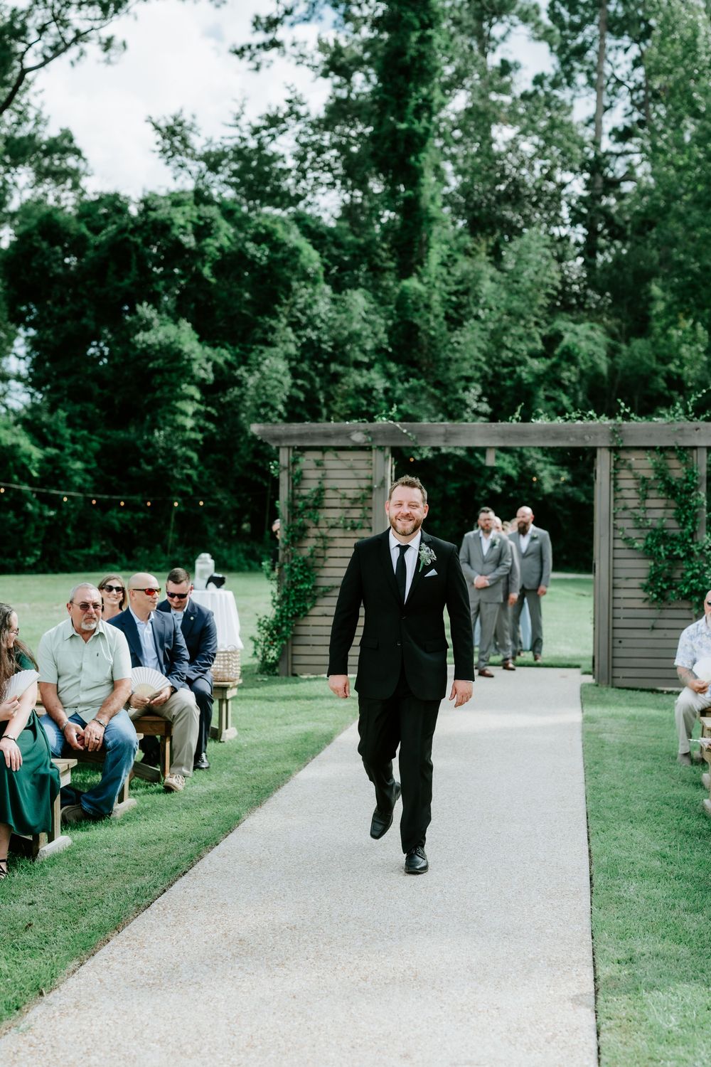 Groom in black suit walks down outdoor wedding aisle lined with seated guests and lush green trees.