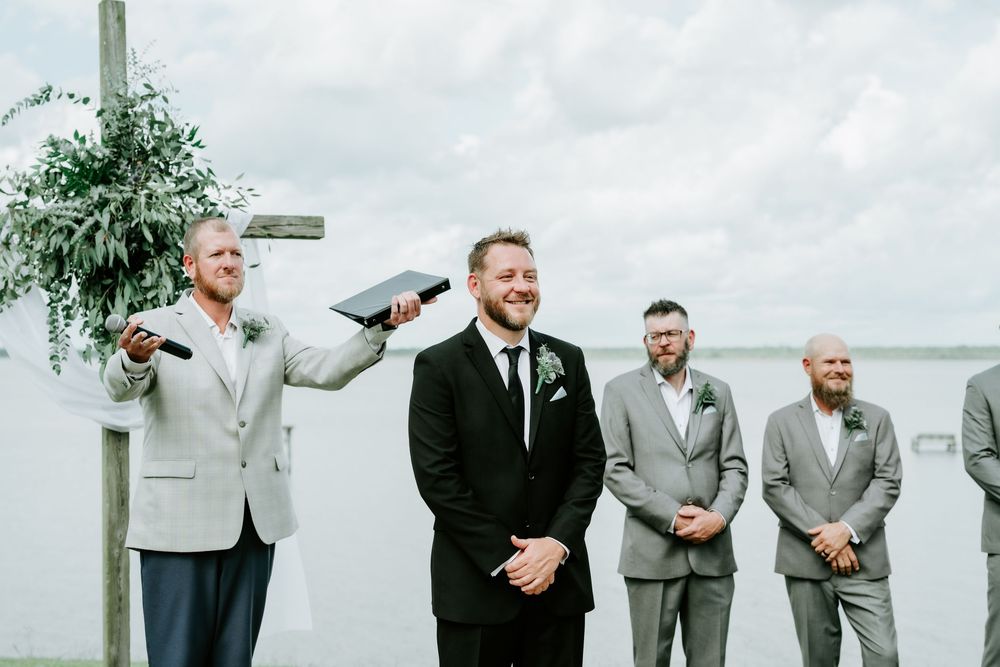 Groom smiling at altar during outdoor waterfront wedding ceremony with groomsmen in gray suits standing nearby.