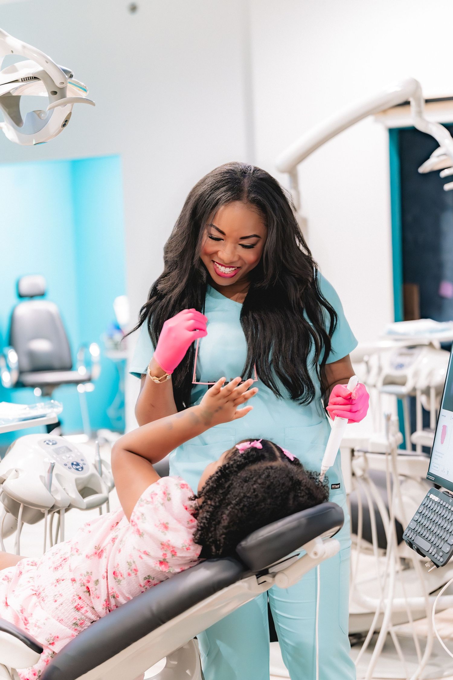 A smiling female dentist in teal scrubs and pink gloves examines a young patient reclining in a dental chair.