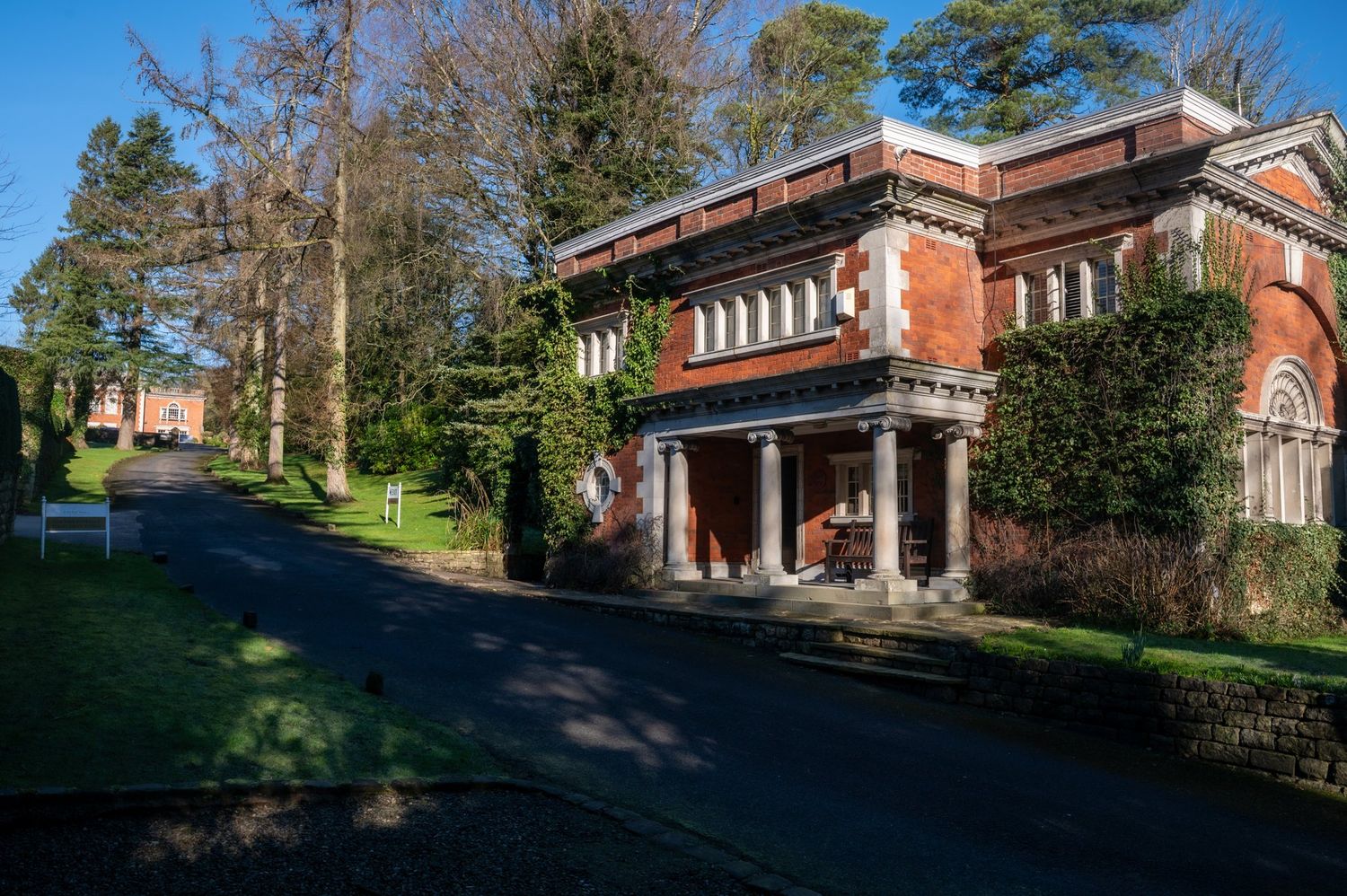 Historic red brick building with classical columns along a tree-lined driveway on a sunny winter day.