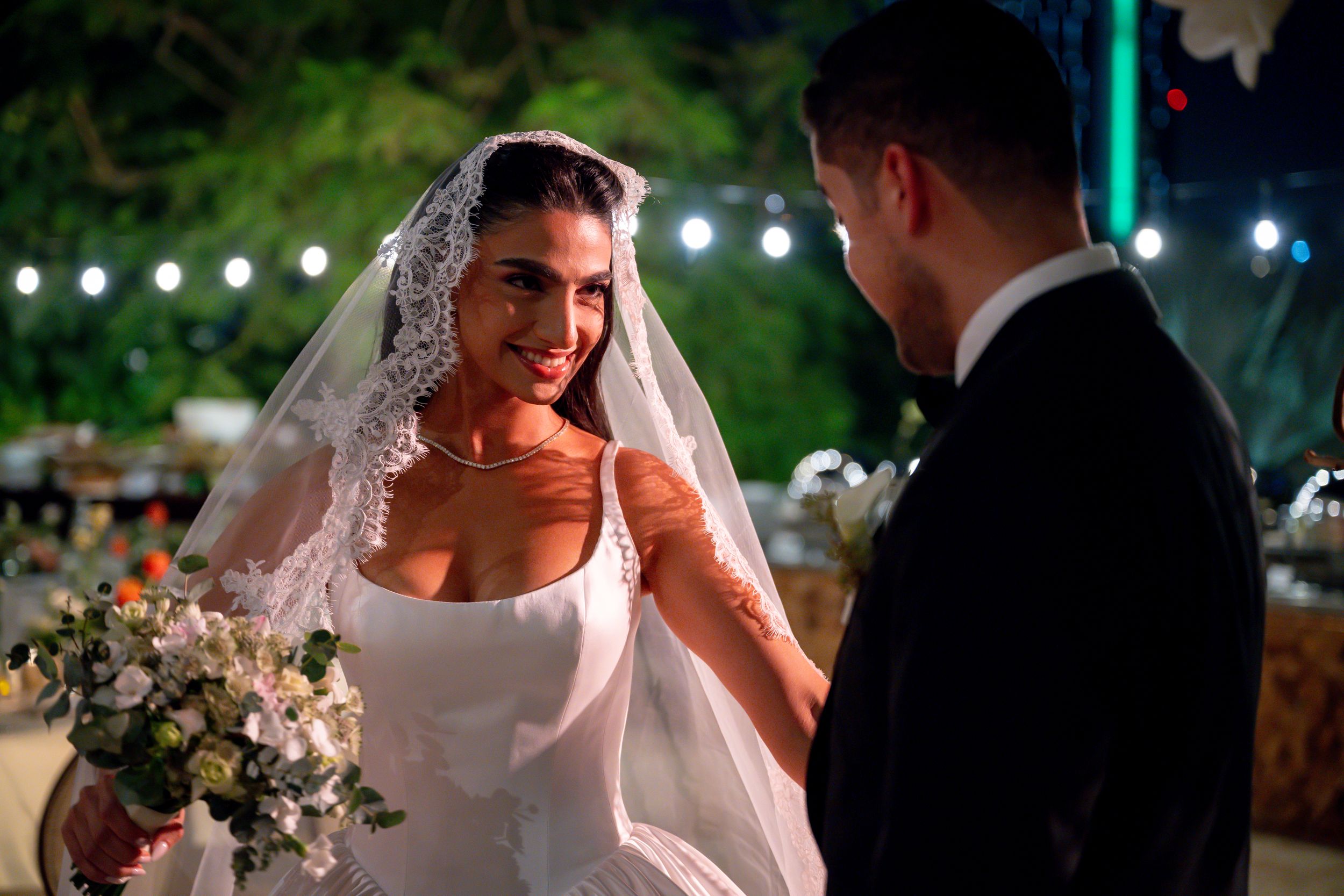 Bride in white gown and veil smiling at groom during outdoor evening wedding ceremony.