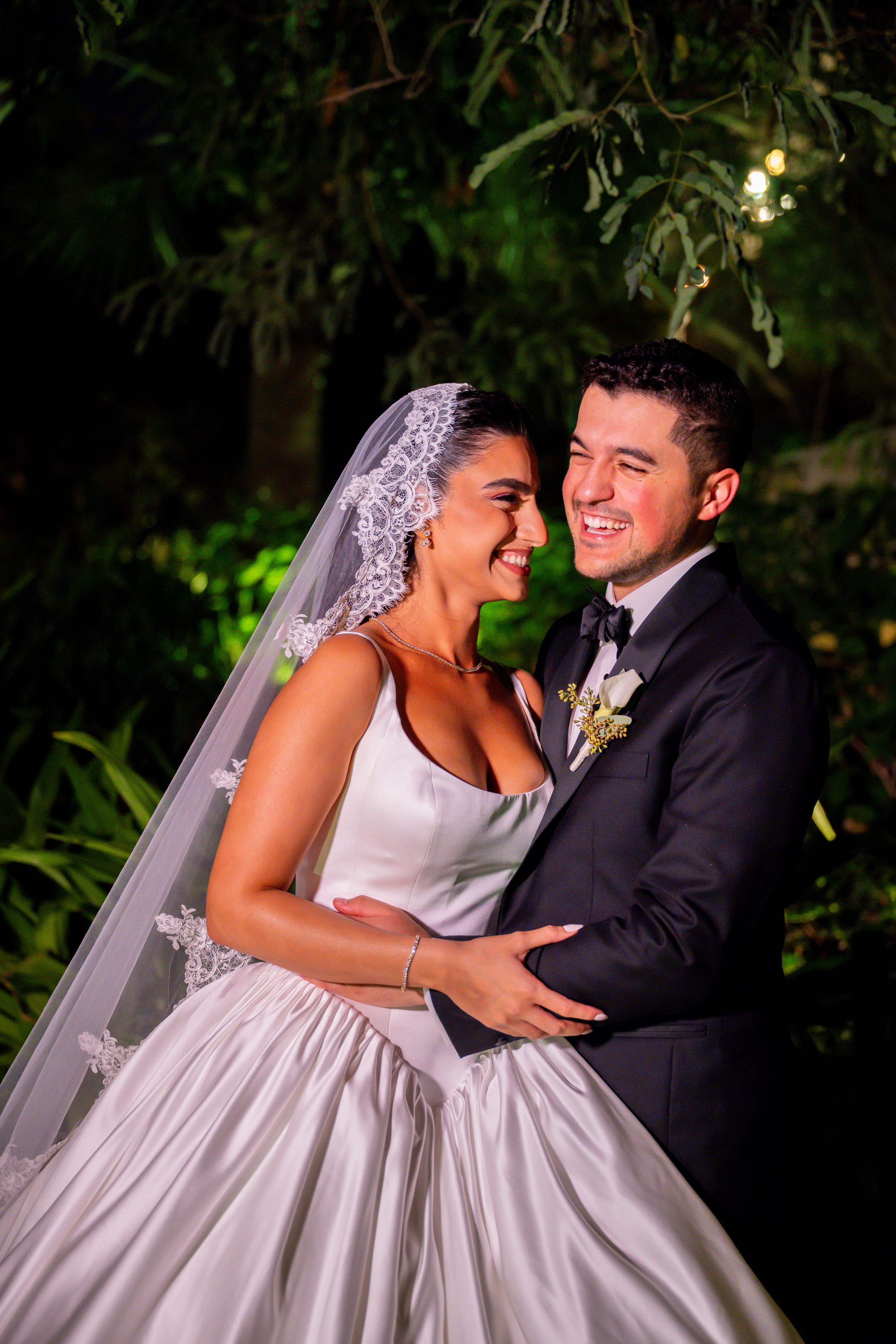 A joyful bride in a white gown and veil embraces her groom in a tuxedo outdoors at night.