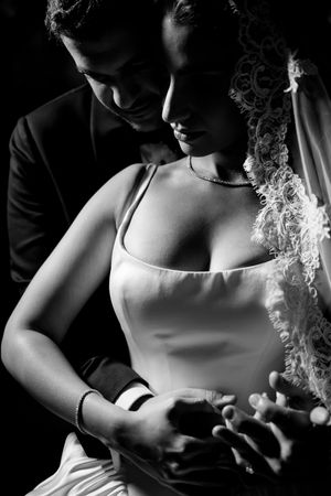 Black and white photo of bride and groom sharing an intimate moment, holding hands in elegant wedding attire.