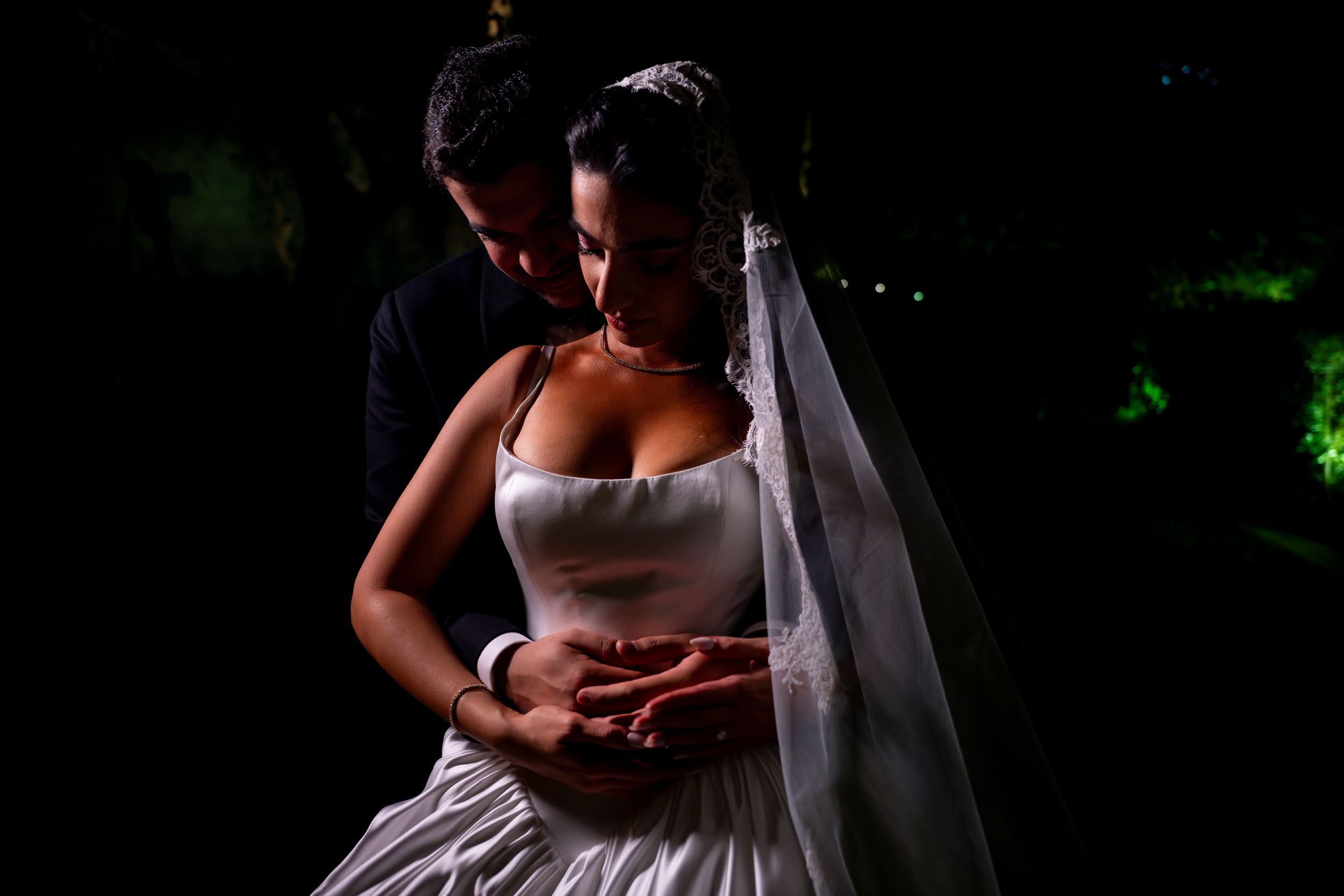 Romantic bride and groom embrace in dramatic dark lighting, her white gown and veil glowing as they hold hands tenderly.
