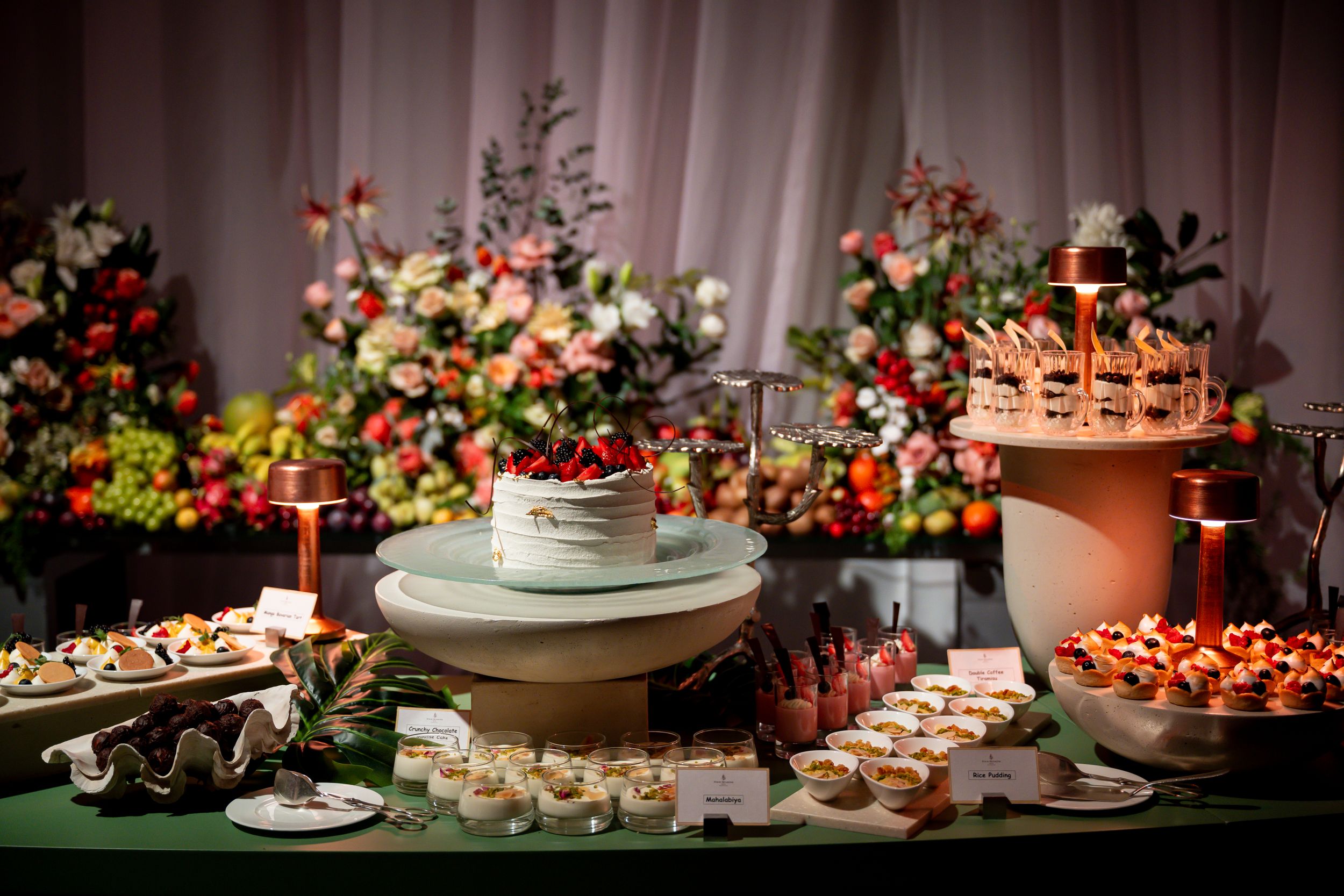 Elegant wedding dessert table with tiered cake, fresh fruit, and floral arrangements on a green tablecloth.