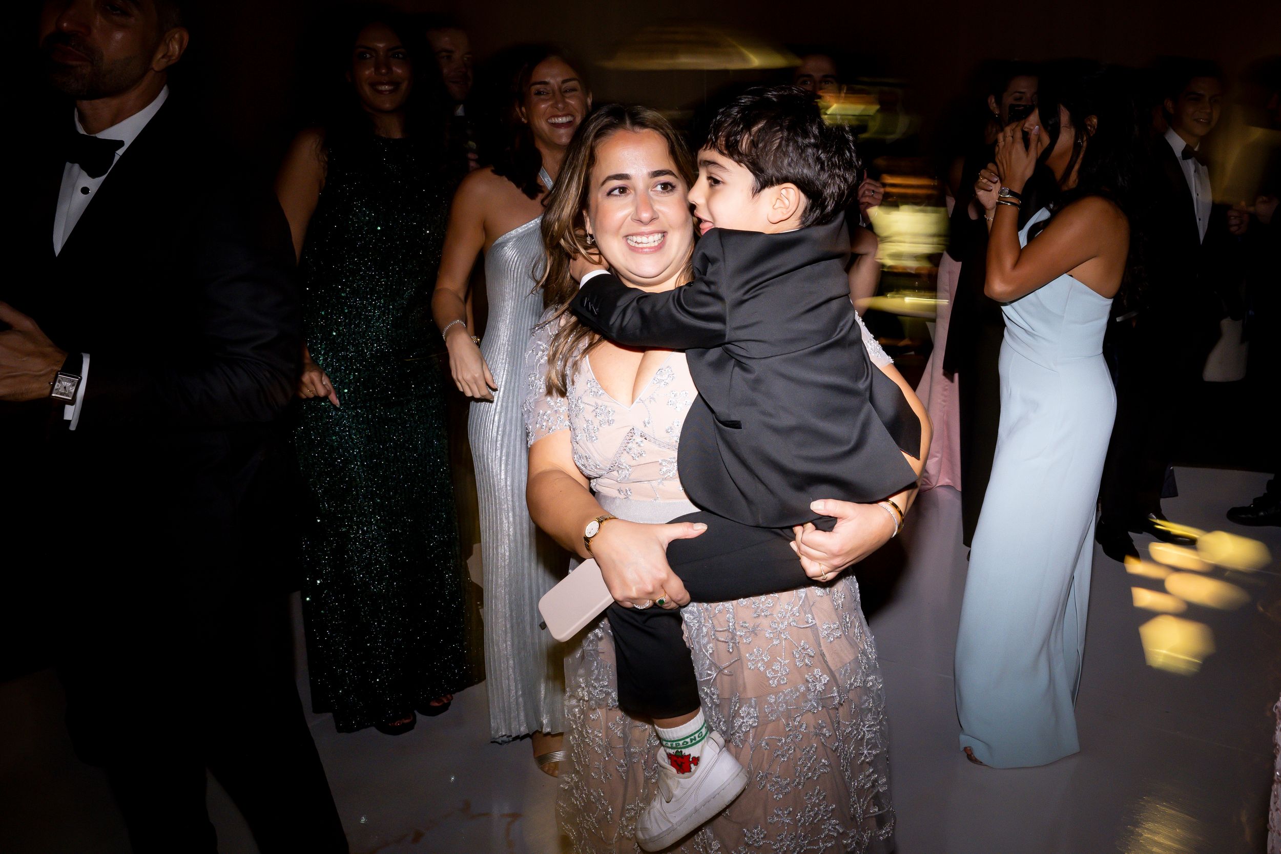 Woman in lace dress holds young boy in suit on a wedding reception dance floor with guests in background.