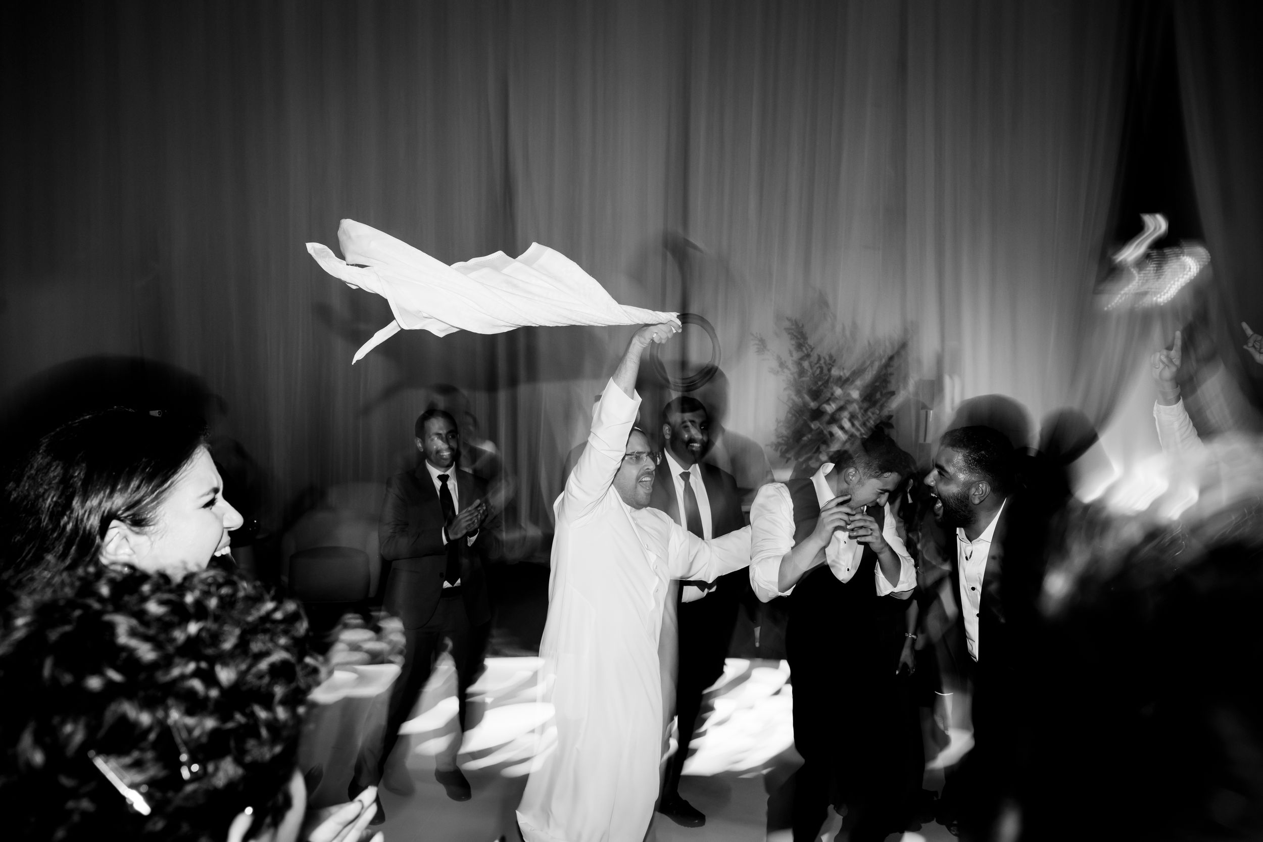 Black and white wedding reception photo of bride dancing joyfully, waving white fabric among celebrating guests.