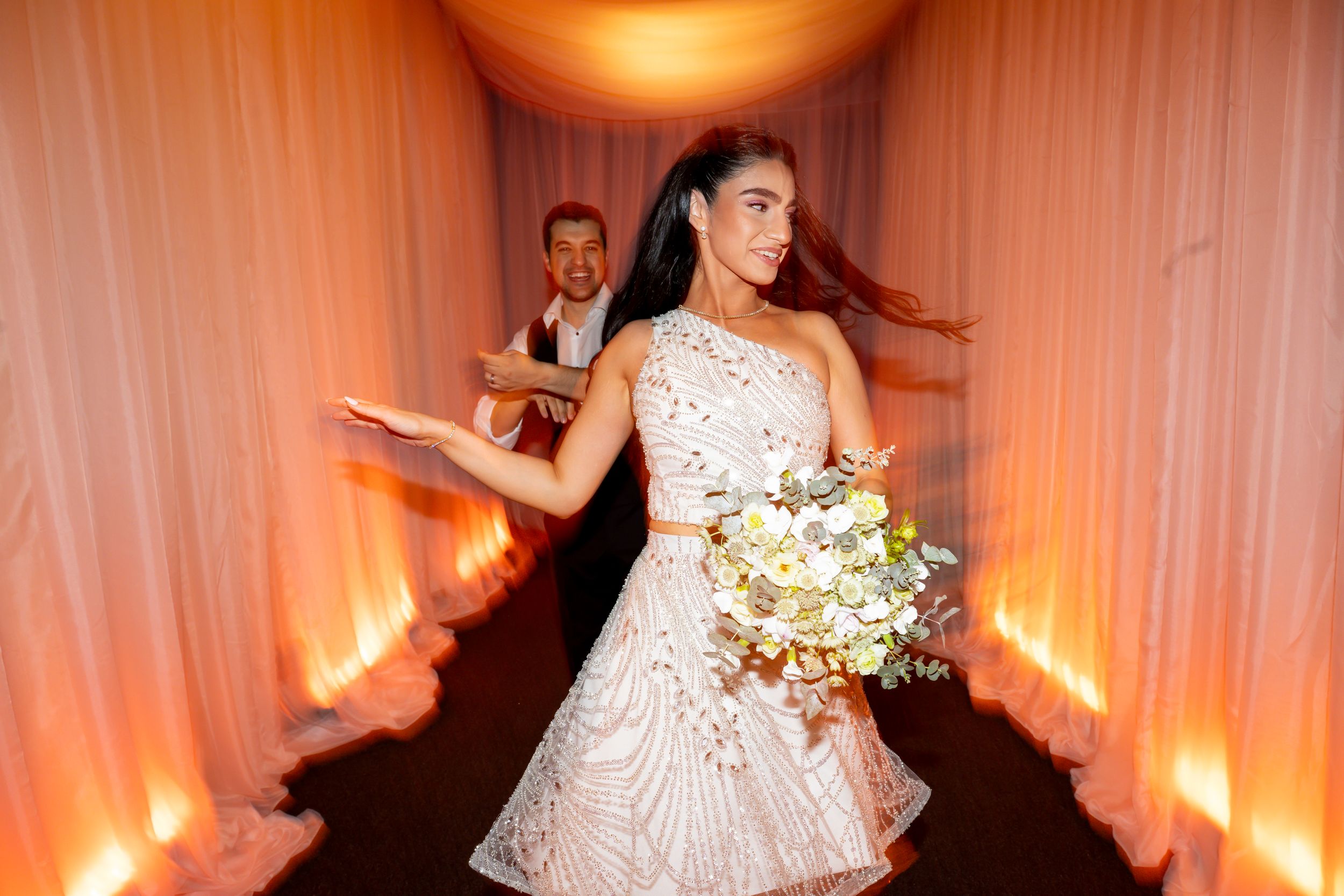 Bride in embellished white mini dress holding bouquet walks through glowing corridor with groom behind her.