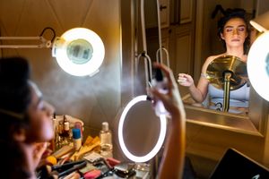 Woman applying makeup at a vanity mirror with ring light and magnifying lamp in a glamorous dressing room.