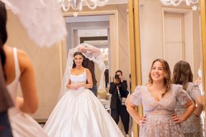 Bride in white ball gown and veil poses before mirror while attendant and photographer capture the moment.